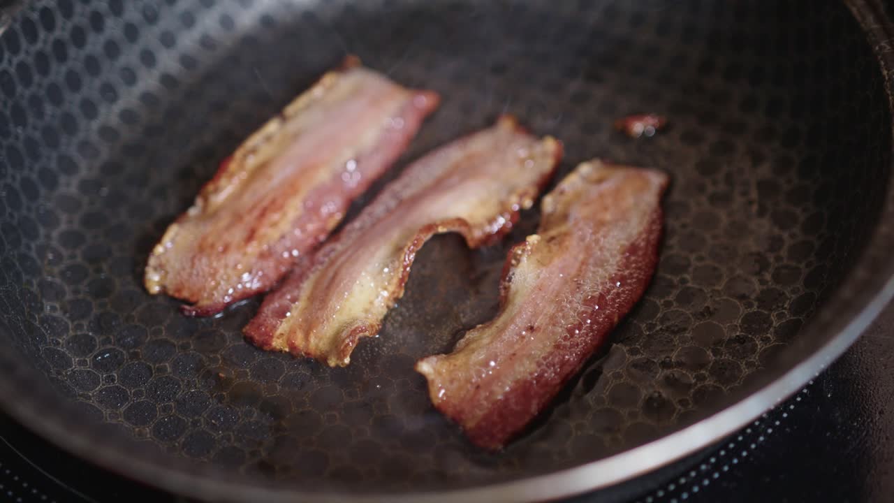Slices of bacon with crisp edges sizzling in a dark patterned frying pan with steam rising. Close up static view