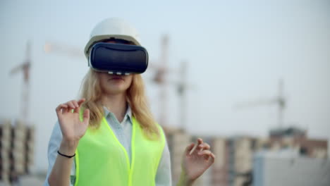 Eine-Designerin-Auf-Einer-Baustelle-Mit-Schutzhelm-Und-Weste-Und-Einer-Virtual-Reality-Brille-Bewegt-Ihre-Hände-Und-Ahmt-So-Die-Benutzeroberfläche-Vor-Dem-Hintergrund-Der-Kräne-Bei-Sonnenuntergang-Nach.