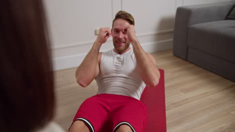 over the shoulder of a happy brunette girl helping her boyfriend with an athletic build with brown hair and stubble in a white t-shirt doing exercises to develop abdominal muscles in a modern apartment during a warm-up and workout session in the morning