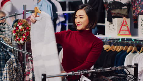asian customer browsing through rack of clothes in christmas themed store, checking for fitting garment size. woman in xmas adorn fashion boutique during winter holiday season