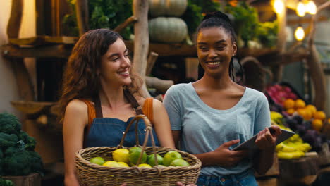 portrait of two smiling women with digital tablet working at fresh fruit and vegetable stall in market