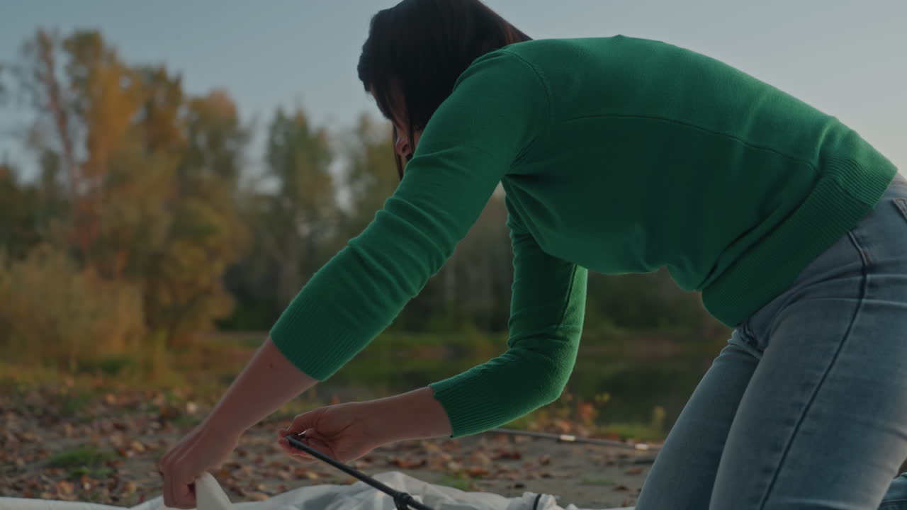 Mujer negra armando una tienda de campaña sola. Mujer afroamericana construyendo un refugio junto al río al anochecer. Mujer independiente con suéter verde arreglando los postes de la tienda al atardecer en la orilla rocosa del río.