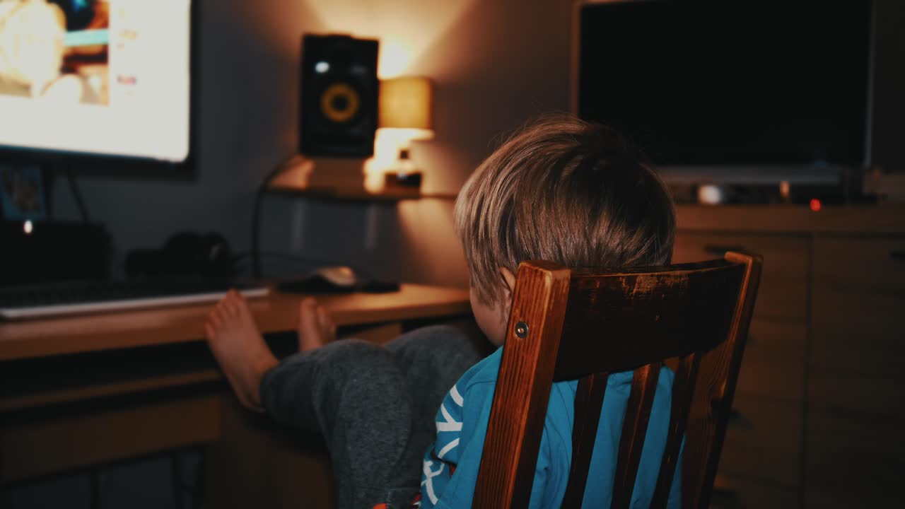 A young boy sits alone in a dimly lit room, gently rocking in a wooden chair while watching cartoons on a monitor. The cozy scene captures a quiet moment of childhood in the evening.
