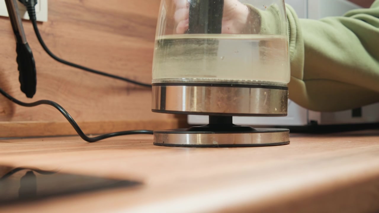 Ahşap tezgahın üzerinde, mutfak aletinin yanında duran yuvarlak metal tabandan kaldırılan cam elektrikli su ısıtıcısının alçak açılı yakın çekimi; hafif bir boşluk, görünür su seviyesi, kablo, yansımalar ve sıcak iç mekan görülüyor.