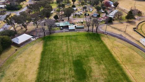 drone camera smoothly ascends above a green sports field bordered by dry grass, revealing surrounding residential neighborhood under natural daylight with steady, wide aerial perspective