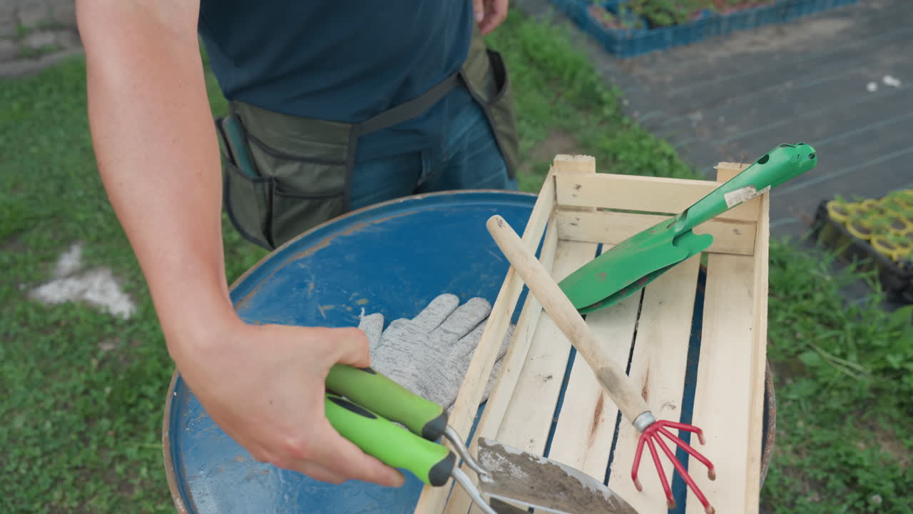 Adult gardener wearing pocket apron removes work tools from pockets and drops gloves onto blue metal barrel beside wooden crate with green trowel and red hand rake