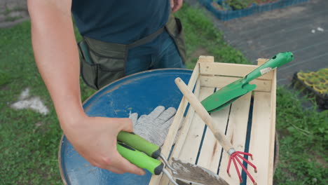 adult gardener wearing pocket apron removes work tools from pockets and drops gloves onto blue metal barrel beside wooden crate with green trowel and red hand rake