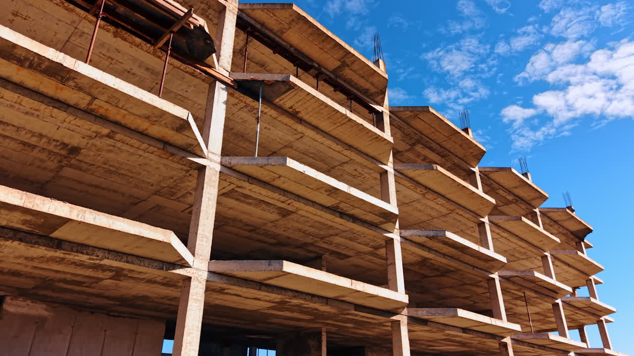Vista de una estructura de hormigón incompleta vista desde abajo, capturando los diversos niveles del edificio. El cielo nublado contrasta con las texturas crudas del hormigón.