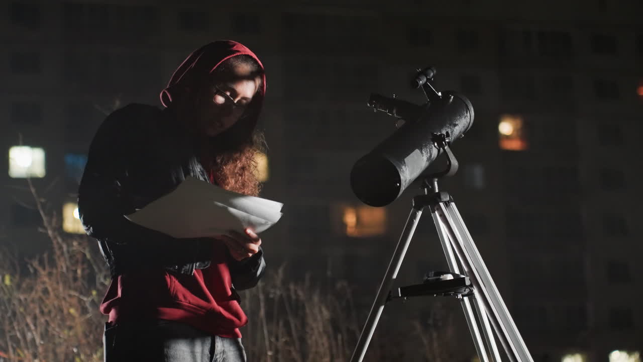 Skjeggete, hvit astronom studerer stjernekart med teleskop, kommenterer loggboken og justerer monteringen, forbereder kamera og okular, lys fra byens silhuett i bakgrunnen, bevisste fokuserte bevegelser