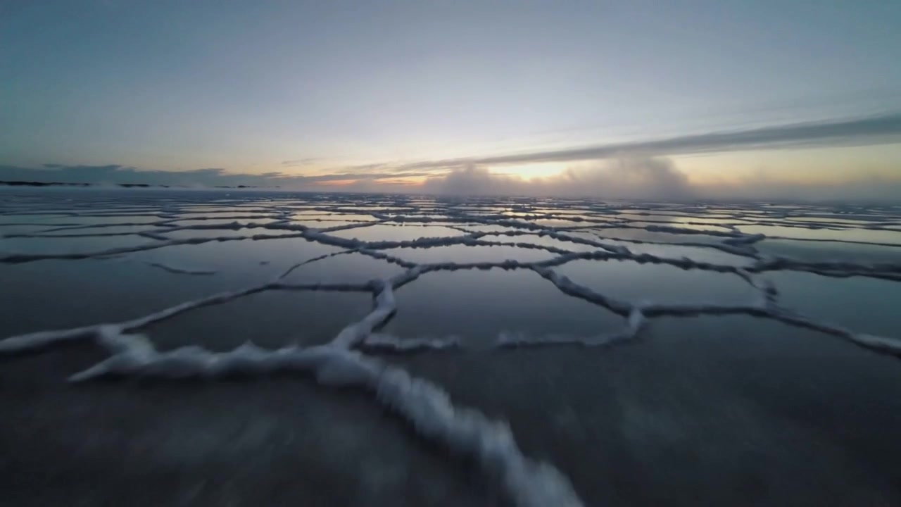 Starting forward glide low camera skimming tidal flat, capturing foam polygon cells and sun glow