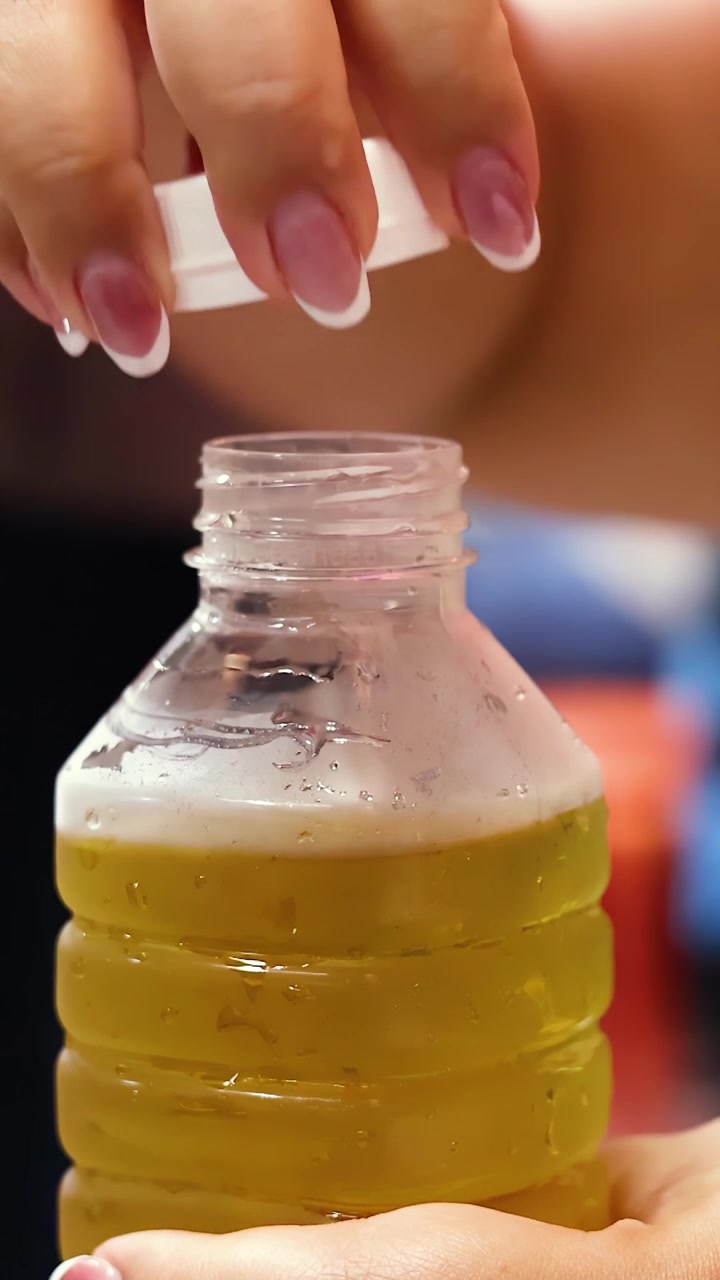Primer plano de una mano desenroscando la tapa de una botella de bebida amarilla fría, en un mercado interior con iluminación suave