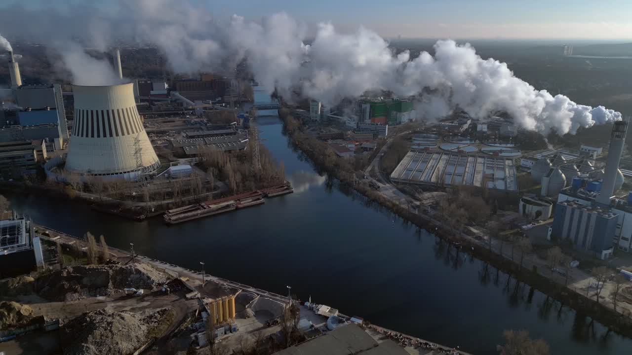 large industrial power plant emitting large amounts of smoke over the city Berlin next to a river on a sunny day. Gorgeous aerial view flight descending drone