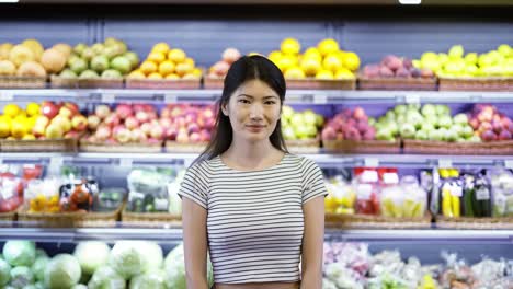 Porträt-Einer-Attraktiven-Jungen-Asiatischen-Frau,-Die-Im-Supermarkt-Mit-Regalen-Voller-Früchte-Im-Hintergrund-Steht,-In-Die-Kamera-Blickt-Und-Lächelt.-Handelsgeschäft-Und-Personenkonzept