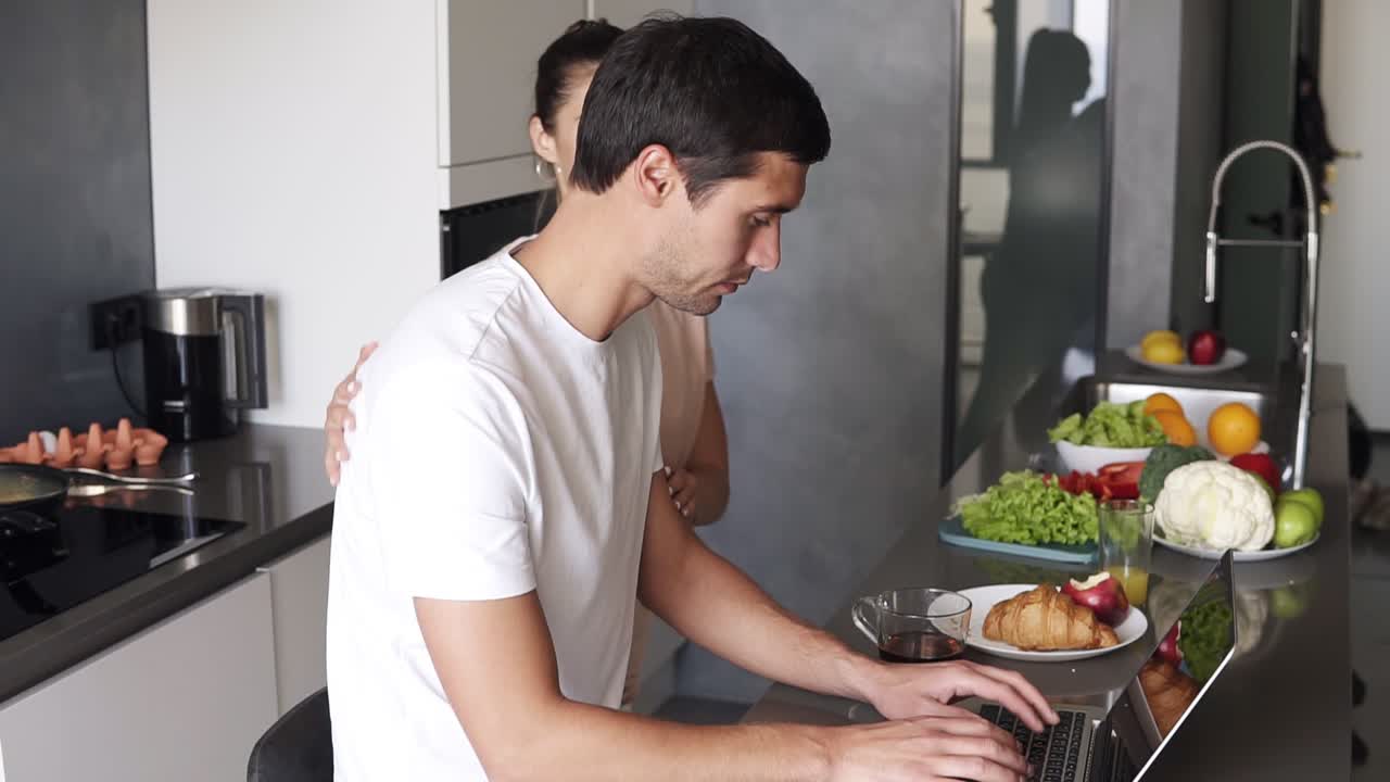 예쁜 젊은 남자는 집에서 주방 카운터에서 아침에 노트북 작업을 앉아서 사용합니다. 남자는 전자 메일, 소셜 미디어 인터넷을 확인하고 키보드에 타이핑합니다. 그의 사랑하는 아내는 그를 키스하고 좋은 아침을 기원합니다. 측면 보기