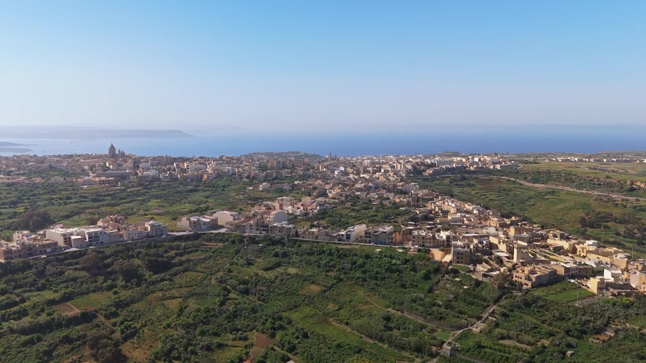 À Nadur, sur l'île de Gozo, une vue aérienne capture les bâtiments résidentiels de la ville côtière, l'architecture maltaise traditionnelle, les terres agricoles et les champs environnants sous un ciel méditerranéen clair
