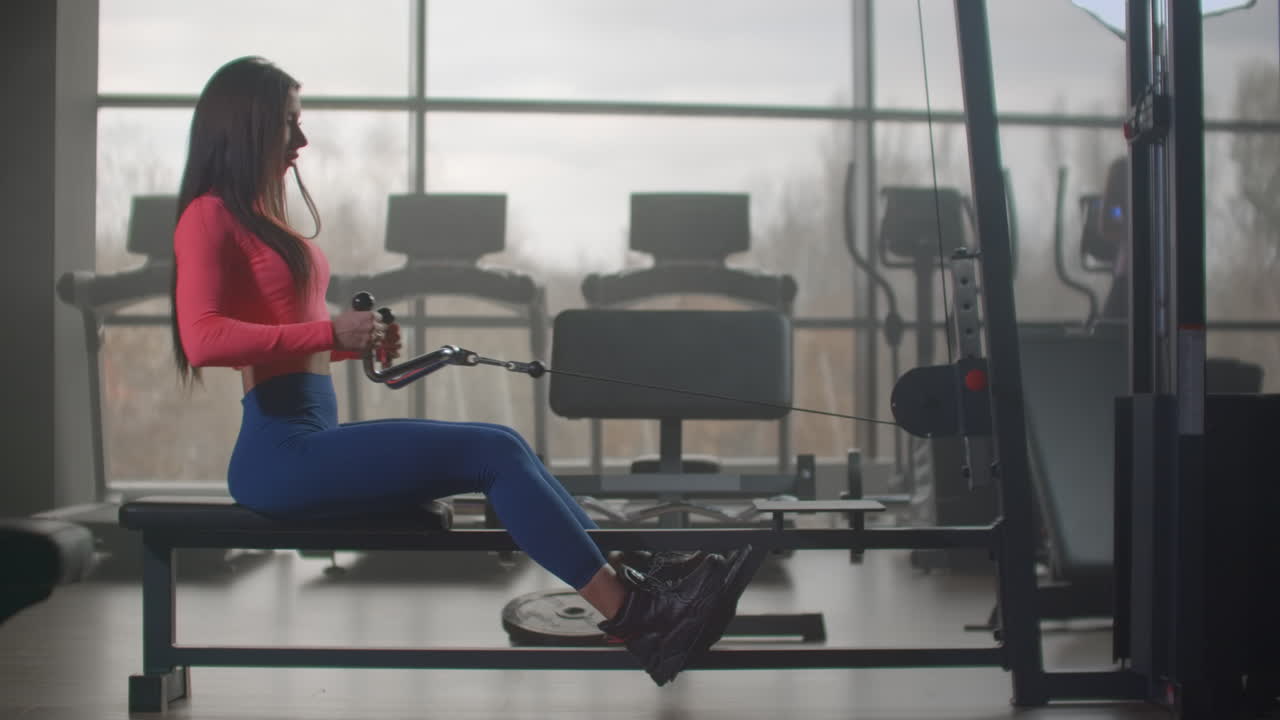 The young female pumps her back and shoulder blades on the training machine. She pulls the handle to the lower abdomen on exhalation and returns to its original position on inhalation.