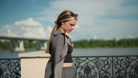 lady in open grey top and black crop top walks near riverfront, passing by a decorative metal fence, background features blurred view of bridge, greenery, while she gazes towards the river