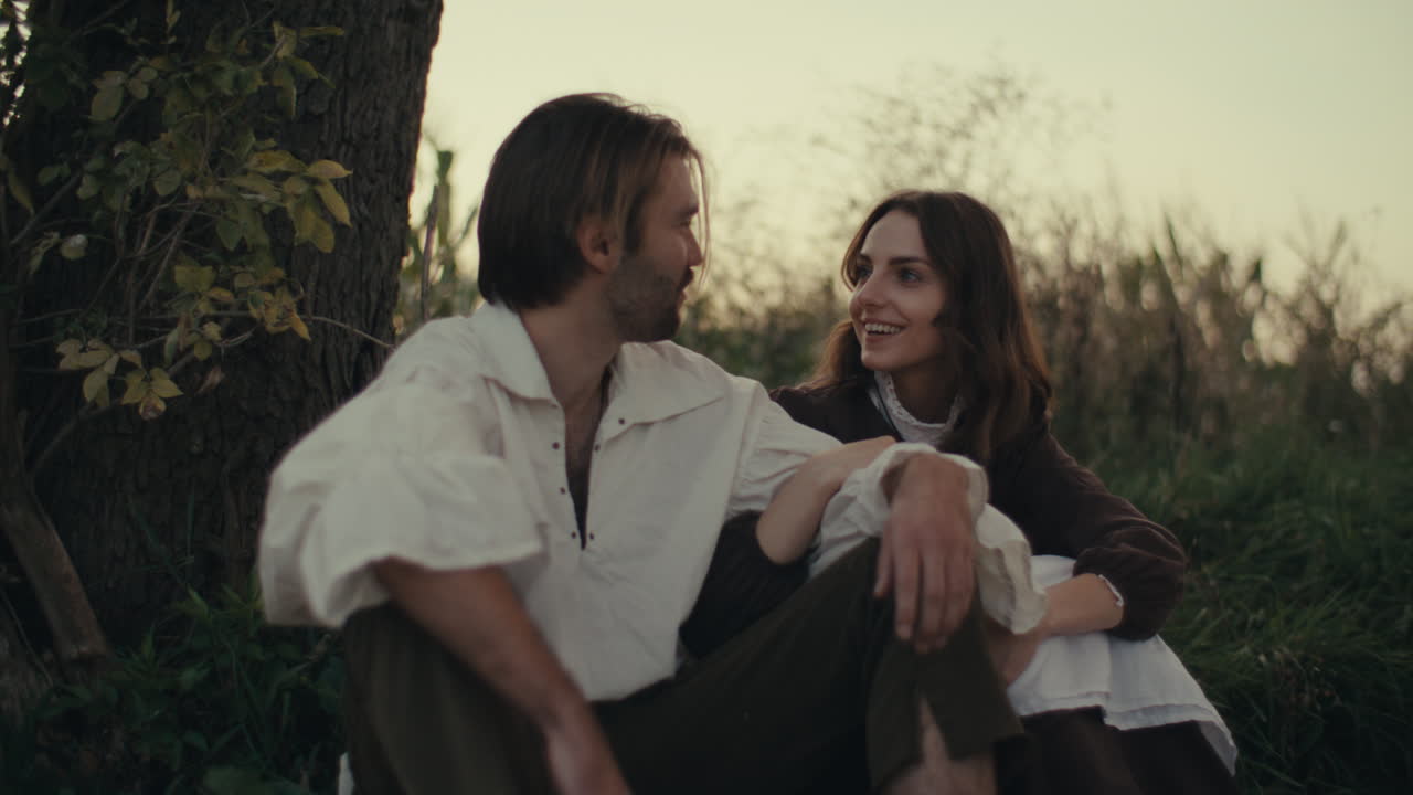 A couple in period costumes sits by a tree in an outdoor setting