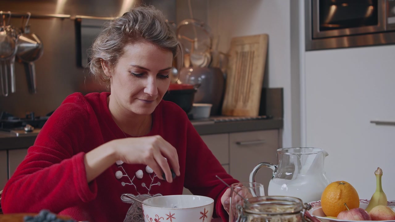 Giovane donna che fa colazione al mattino bevendo tè caldo e mettendo mirtilli nel suo yogurt. Indossa un maglione natalizio festivo, felice e che si gode il momento