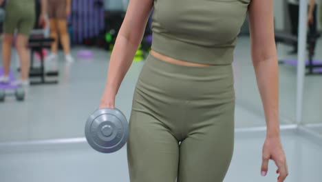 partial view of woman gripping dumbbell while turning around and resting leg on bench, gym environment with various fitness equipment, mirrors, and active setting reflects strength training