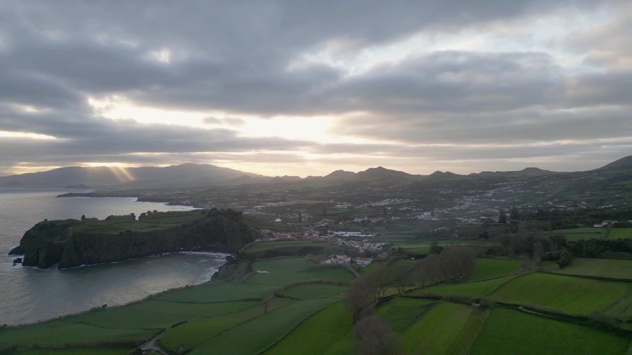 Vista aérea panorâmica de Ponta Delgada nos Açores durante o pôr do sol, com campos verdes exuberantes, falésias costeiras e montanhas distantes sob nuvens dramáticas