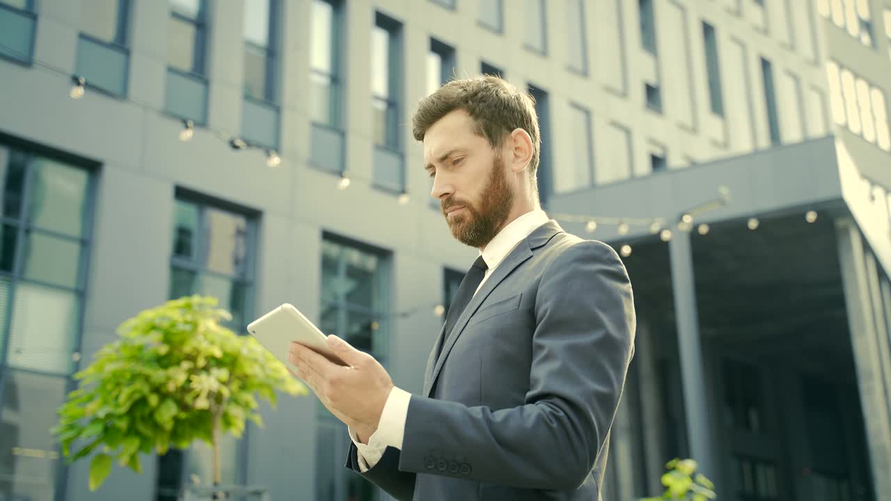elegante hombre de negocios barbudo en traje de negocios formal de pie trabajando con la tableta en las manos en el fondo edificio de oficinas modernas afuera.