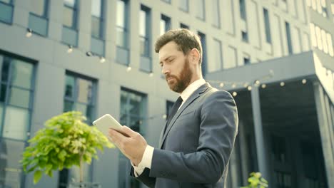 stylish bearded businessman in formal business suit standing working with tablet in hands on background modern office building outside.