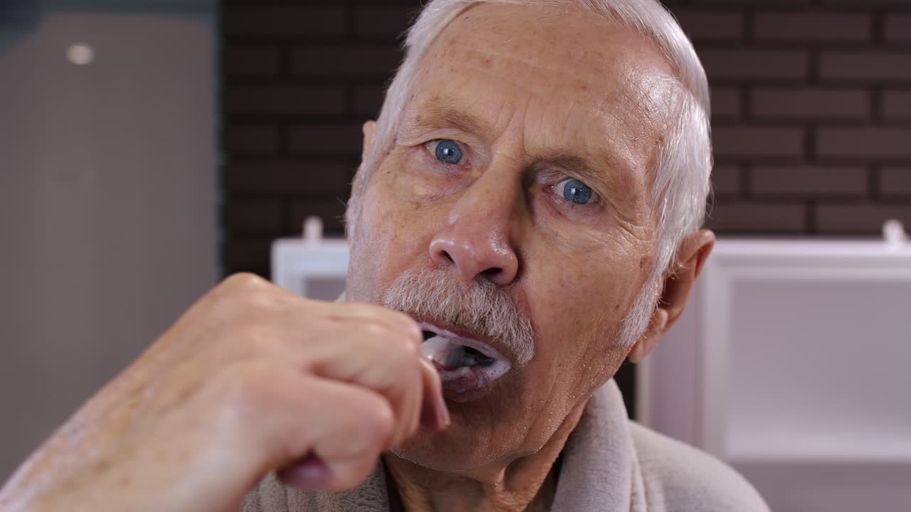 Senior man grandfather in bathrobe brushing teeth, looking into mirror. Morning hygiene at bathroom