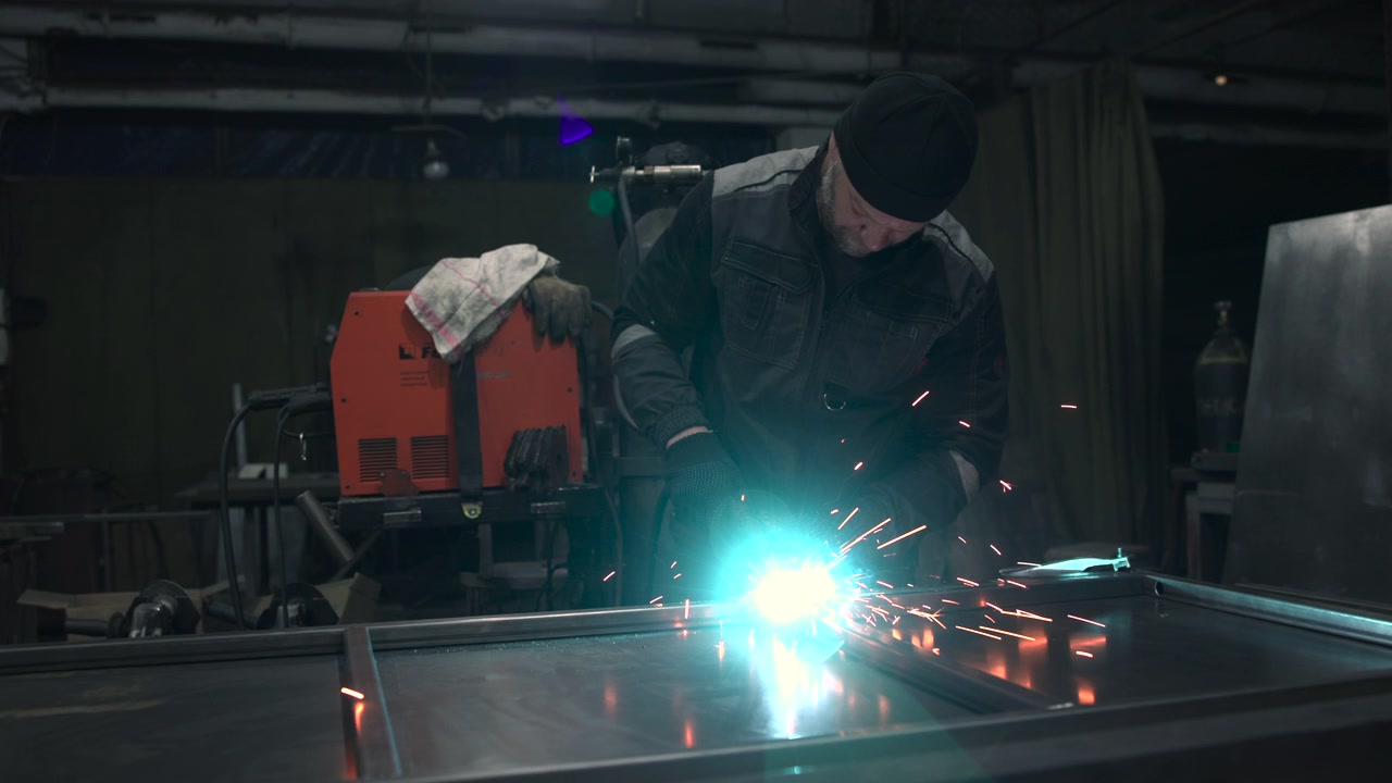 Adult worker in beanie and protective clothing bends over large steel panel while handheld tool sends bright sparks across dim workshop, creating intense industrial atmosphere with precision, heat