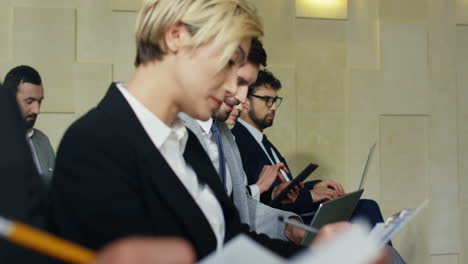 Close-up-view-of-multiethnic-business-people-sitting-on-chairs-in-a-conference-room-and-typing-on-laptops-and-tablets-while-listening