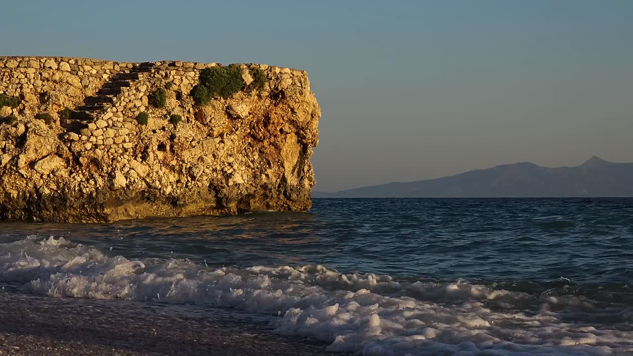 Ancient stone walls of fortress on shoreline, splashed by sea water at sunset golden hour, standing against dark blue sea in Mediterranean
