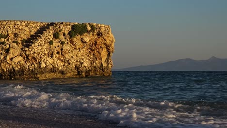 Antiguos-Muros-De-Piedra-De-La-Fortaleza-En-La-Costa,-Salpicados-Por-Agua-De-Mar-En-La-Hora-Dorada-Del-Atardecer,-Frente-Al-Mar-Azul-Oscuro-En-El-Mediterráneo