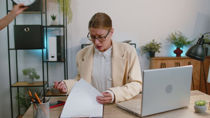 Business woman working on laptop at office with anxiety checking time on clock being late, deadline