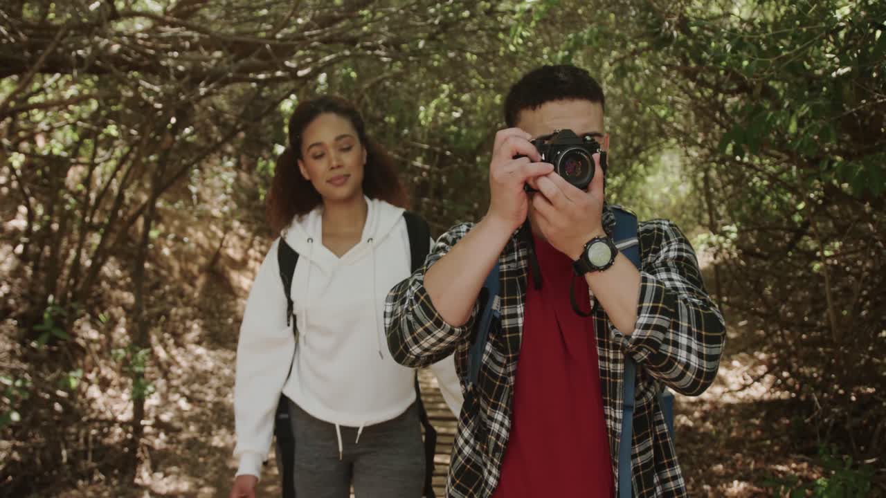 feliz pareja afroamericana con mochila, caminando y tomando fotos en el bosque, cámara lenta