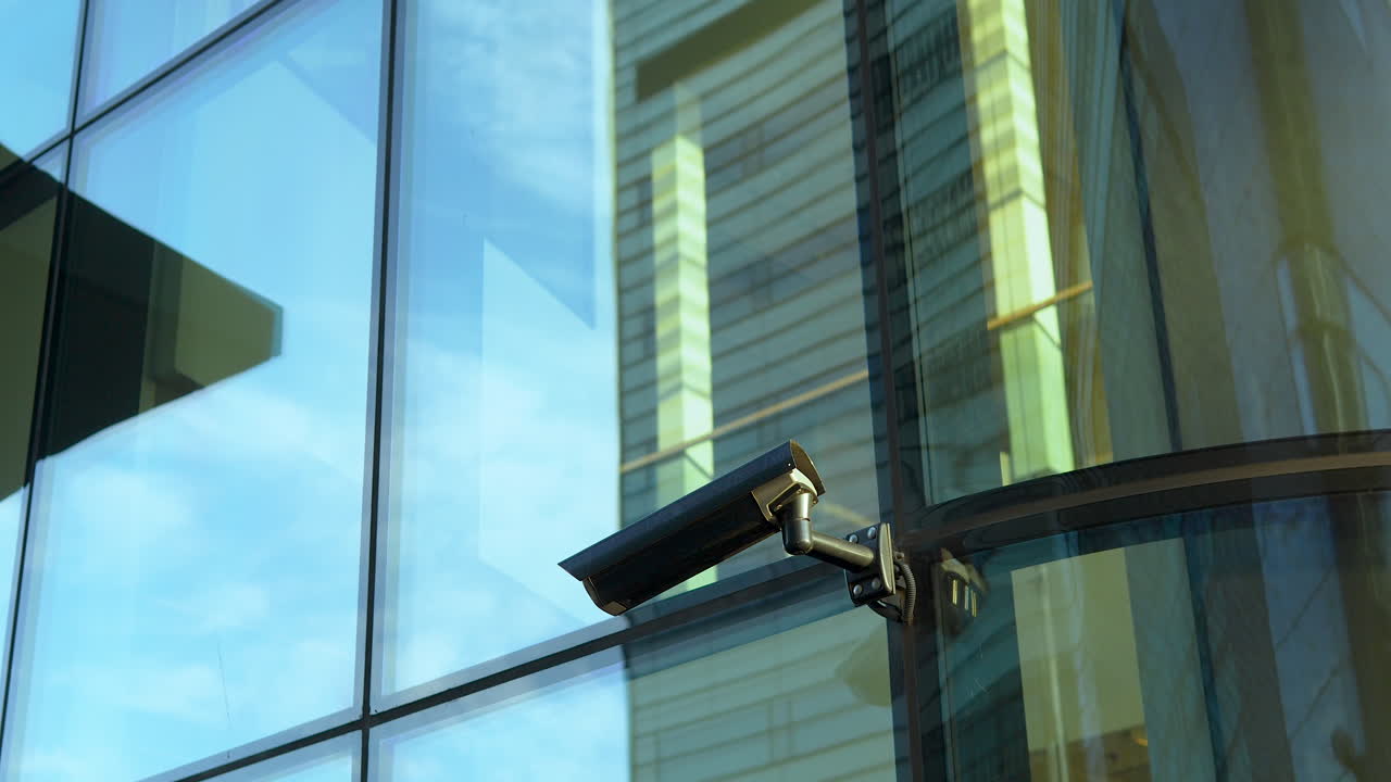 Surveillance camera attached to a modern building's reflective glass facade with the city's distorted reflection