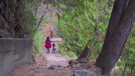 una madre y su hija caminan por un sendero forestal a fines del otoño con hojas por todas partes al lado de un árbol verde y hojas naranjas con un lago