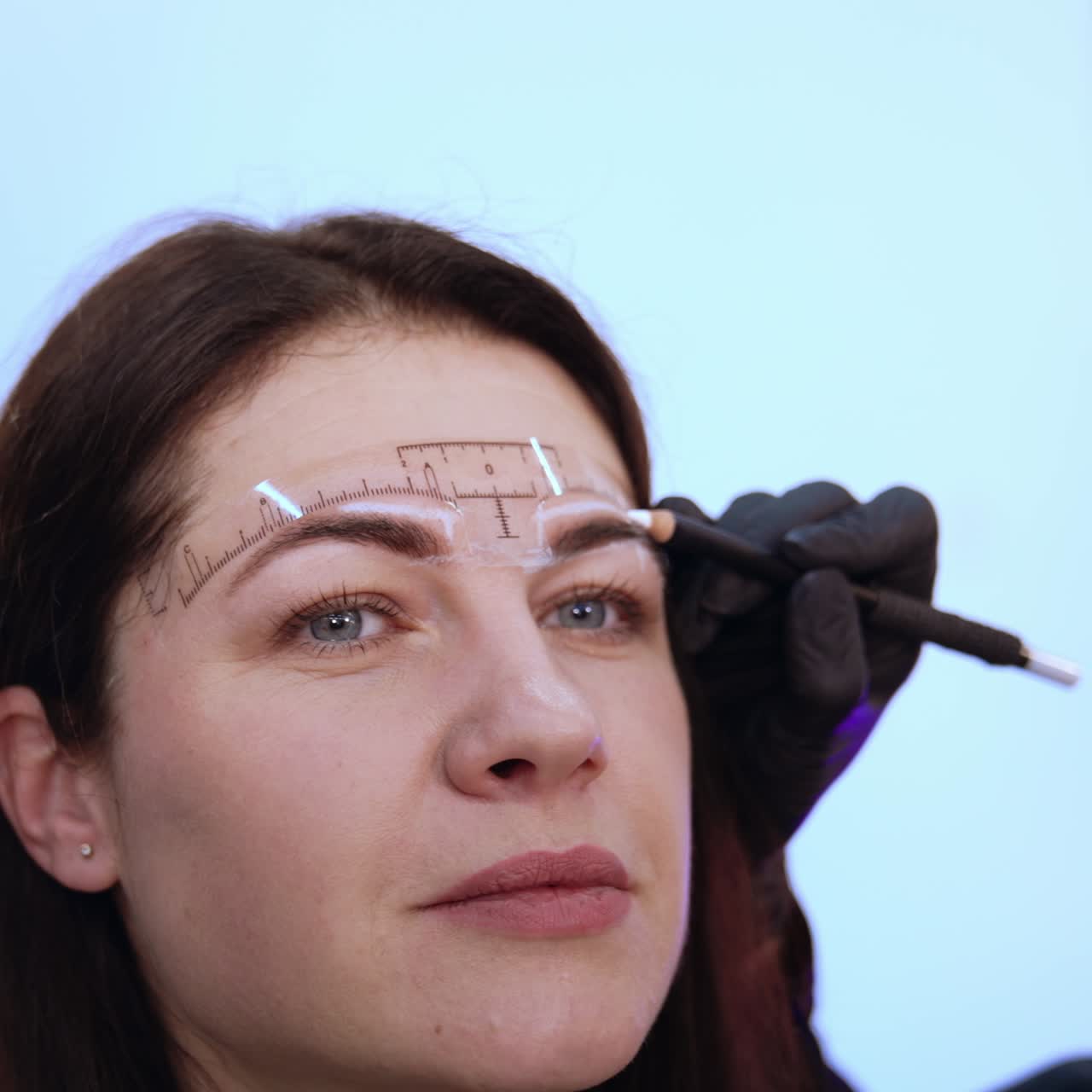 Beautiful adult brunette is having the eyebrow correction procedure. Cosmetologist drawing white lines to mark the limits of eyebrow make up