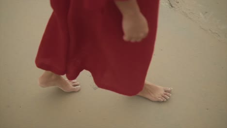 slow motion side view of asian indian young woman wearing beautiful red dress, walking barefoot by beach while wind is blowing the dress in the air- female tourist on summer vacation