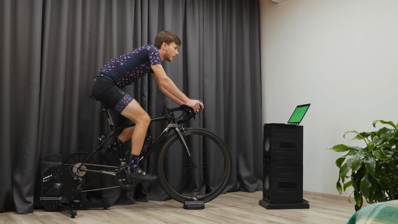 정지 자전거 트레이너에서 심장 훈련을 하는 젊은이. 프로 자전거 선수가 경쟁을 준비하고 컴퓨터의 녹색 화면을 보고 있습니다.