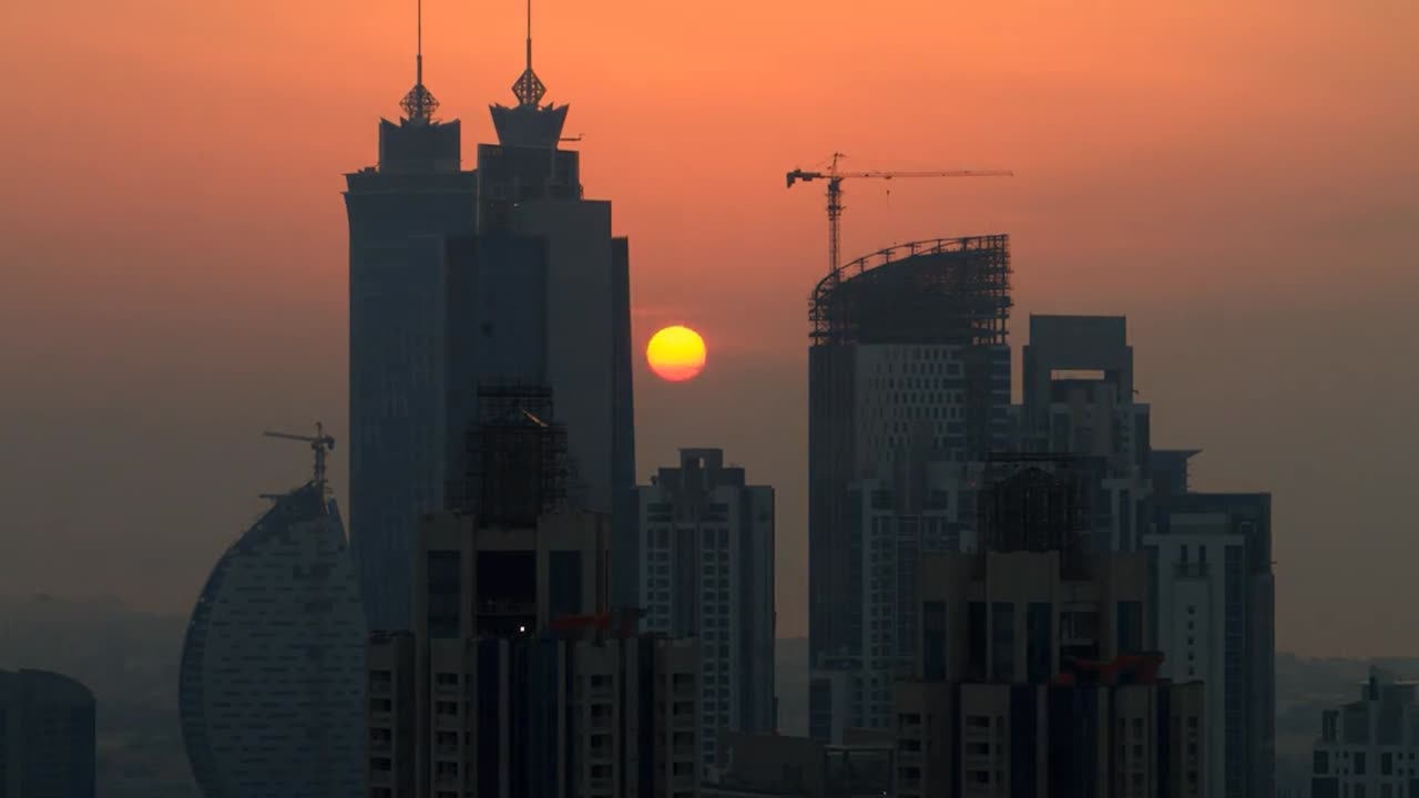 Metraje en time-lapse del horizonte de una ciudad moderna al atardecer, con el sol parcialmente oculto por un alto rascacielos. La escena muestra edificios de gran altura, algunos en construcción con grúas visibles.
