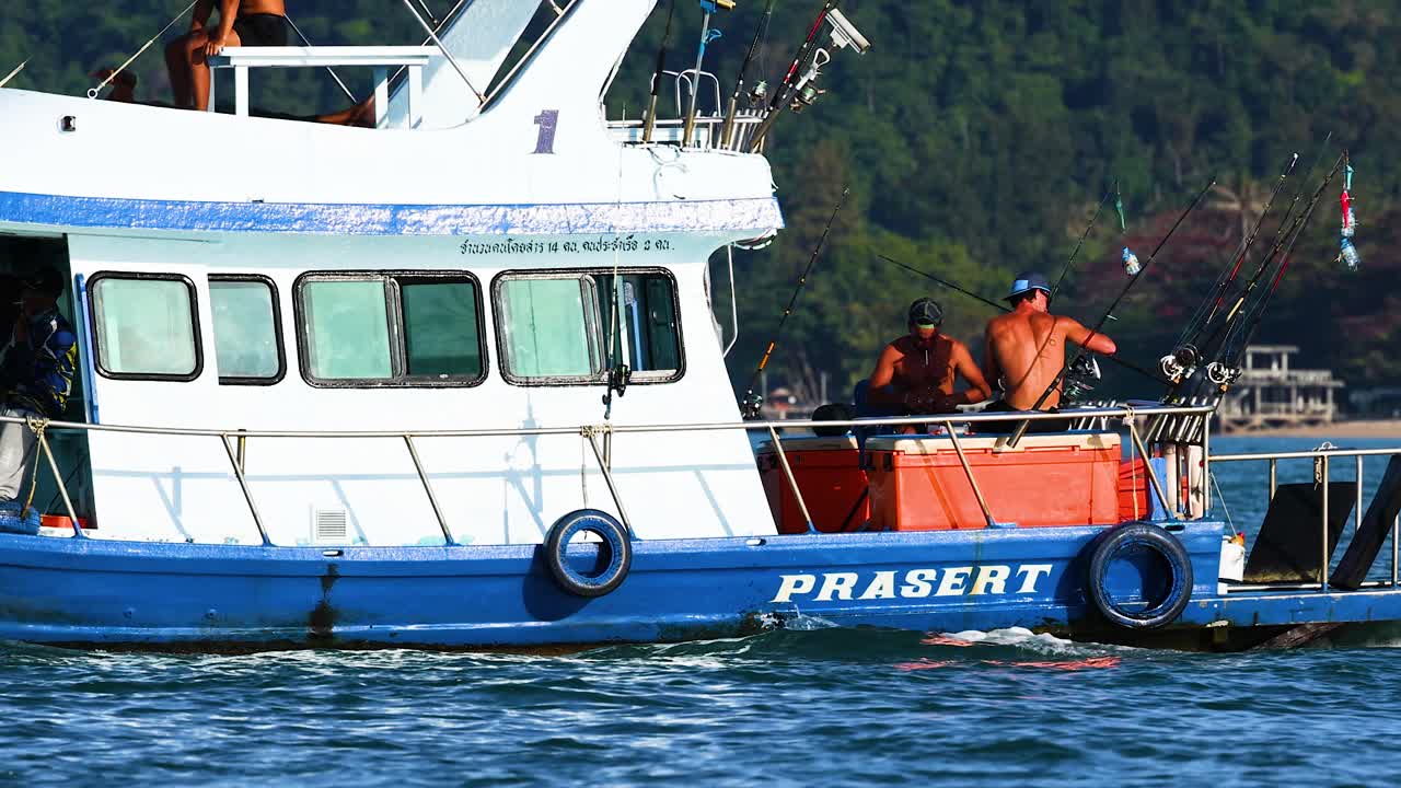 A fishing boat with crew members operates in the waters off Phuket, Thailand, under bright daylight with lush greenery in the background