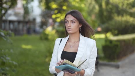 Joven-Mujer-De-Negocios-Caminando-Y-Leyendo-Su-Cuaderno-En-Un-Parque