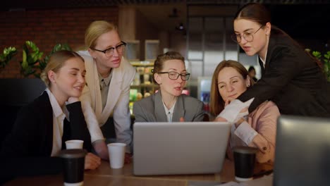 A-confident-middle-aged-blonde-girl-in-glasses-and-a-gray-uniform-communicates-with-her-businesswoman-colleagues-while-sitting-at-a-table-in-front-of-a-laptop-in-a-modern-office