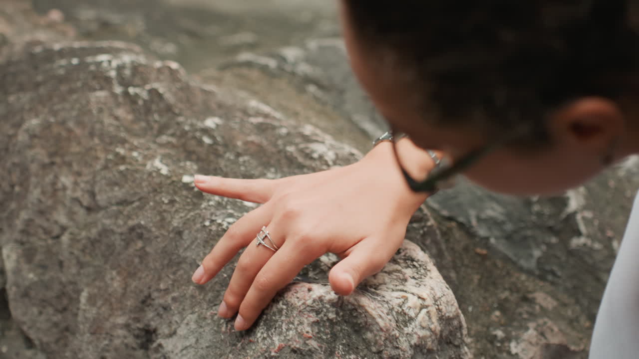 Closeup hands touching weathered granite rock, woman wearing bracelet and rings gently explores textured boulder at seaside, soft overcast light, contemplative mood, tactile curiosity and slow paced