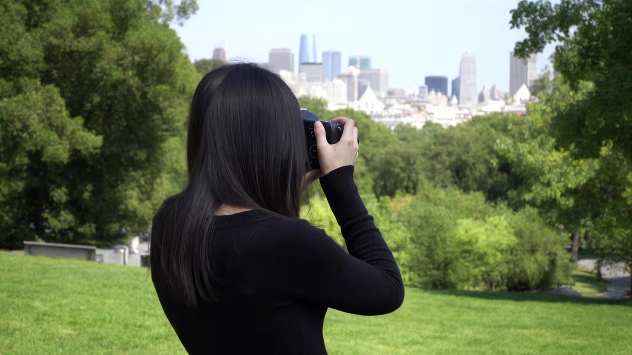 Woman Photographer In Park Framing Cityscape, Back View Capturing Skyline With Camera, Lush Trees And Green