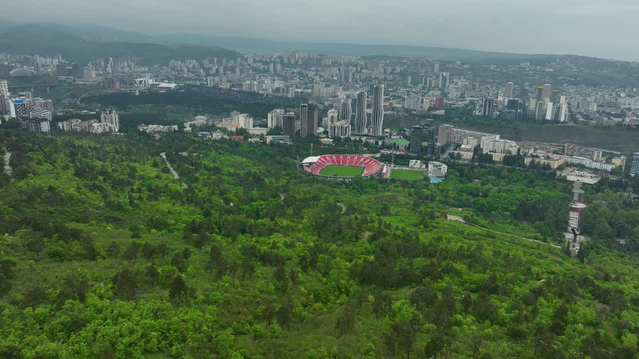 Aerial view of Tbilisi city framed by green hills and the red roof of the Dinamo Arena stadium visible amid the urban landscape