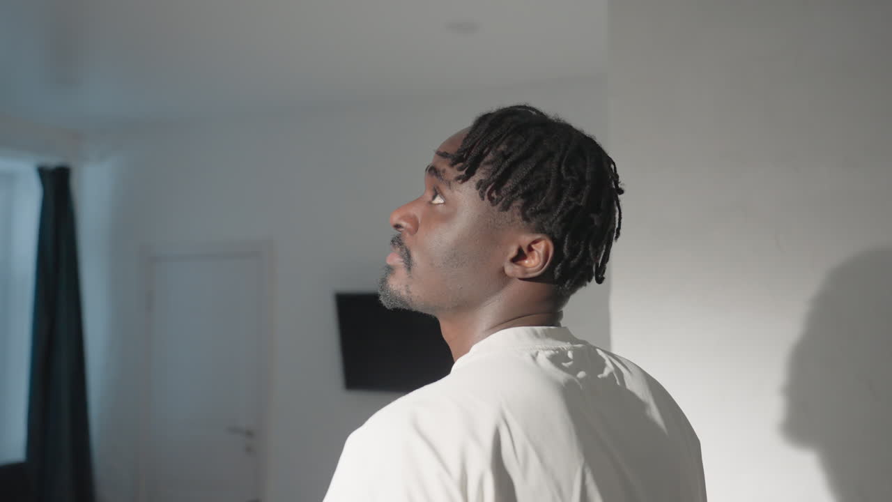Person with short dreadlocks standing in front of a modern, minimalistic living room. The image captures their back, showcasing a simple white t-shirt in a well-lit, serene environment with a TV in the background