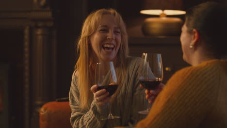 two female friends relaxing and making toast on sofa in lounge with cosy fire holding glass of wine