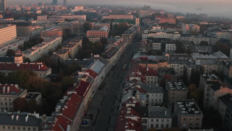 Backwards-fly-above-buildings-in-central-district.-Tilt-up-reveal-of-cityscape-with-buildings-lit-by-rising-sun.-Warsaw,-Poland