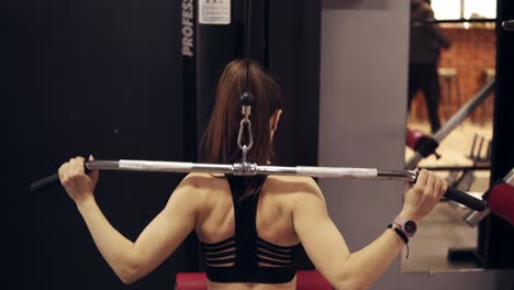 Backside-footage-of-an-athletic-strong-brunette-female-pulling-the-bar-of-lat-pulldown-mashine-at-the-gym.-Getting-body-ready-for-summer.-Achieving-perfect-shapes.